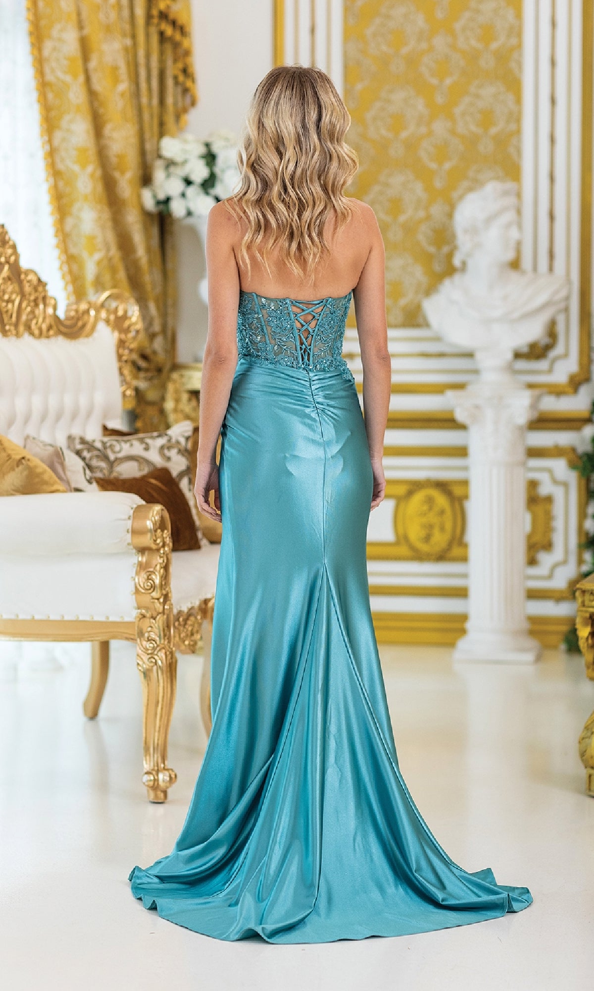 A woman in the Strapless Long Prom Dress: Dancing Queen 4532, featuring a turquoise strapless design with an embellished corset bodice, stands in an ornate, gold-accented room with classical decor and elegant furnishings.